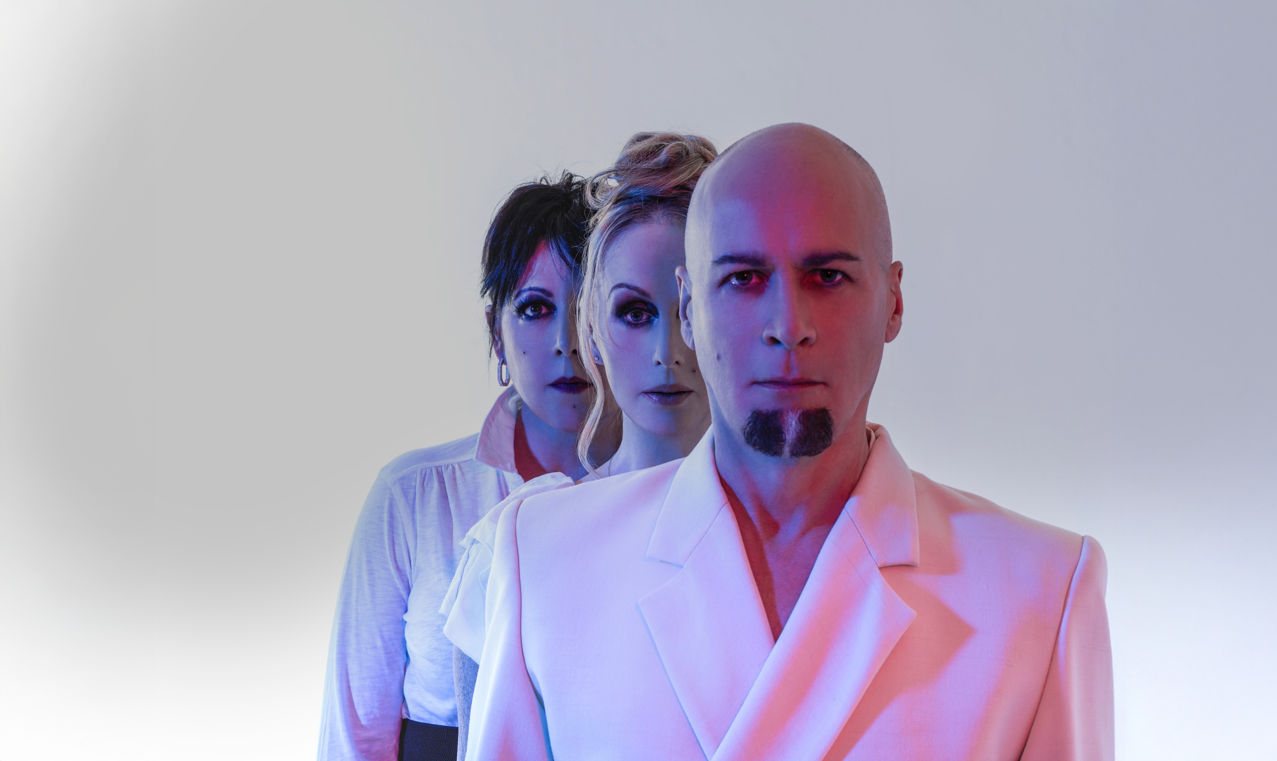 Portrait of 3 members of the band Human League. They are wearing all white and have pink and blue lighting on them.